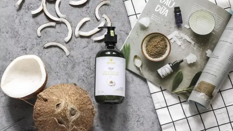 Bottle of argan oil with a pump on a textured gray surface, surrounded by a halved coconut, a small bowl of powder, and an open magazine.