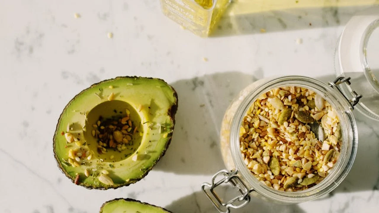 Halved avocado next to a jar of chopped nuts on a light countertop, with a bottle of oil visible in the background.