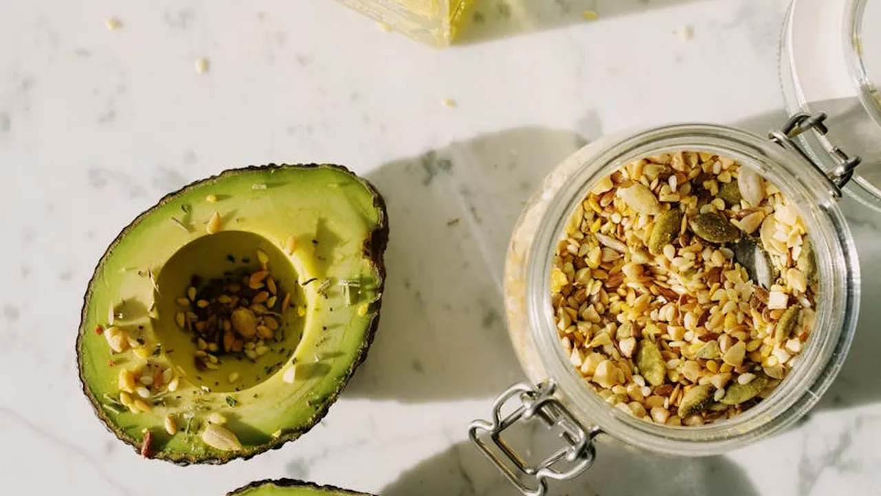 Halved avocado next to a jar of chopped nuts on a marble surface