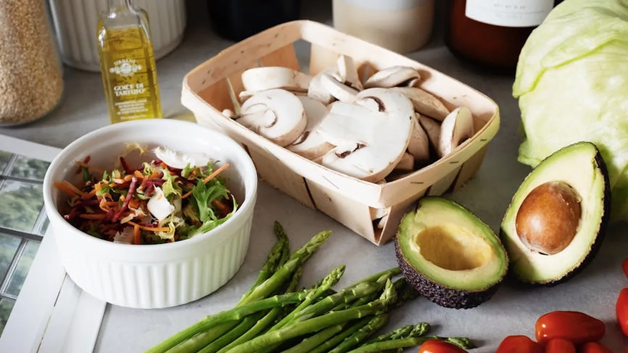 A kitchen counter with fresh produce: avocado halves, mushrooms, asparagus, a bowl of salad, and a bottle of oil.