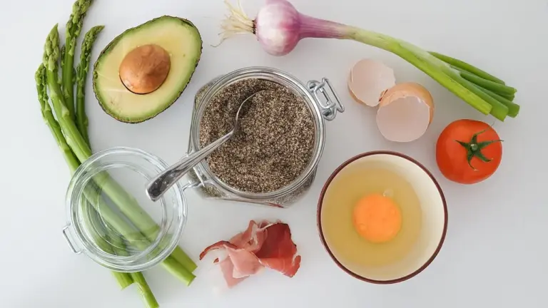 Assorted fresh ingredients on a light surface: avocado half with pit, asparagus, garlic, shallots, green onions, tomato, eggs, and a jar of spices.