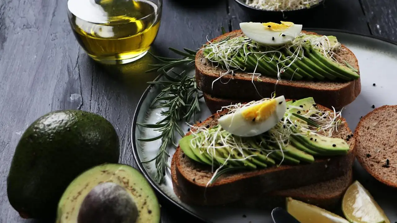 Two slices of whole-grain toast topped with avocado slices, sprouts, and a soft-boiled egg, with a bottle of oil and whole avocados visible in the background.