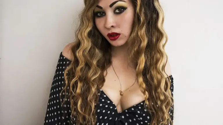 Woman with long, wavy hair wearing an off-shoulder black polka-dot top, posing against a plain background.