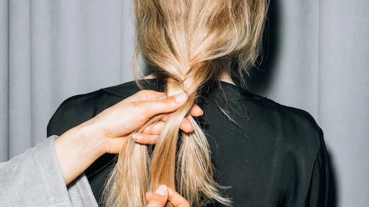 Back view of a person with long blonde hair, a hand gathering a strand as if to tie it.