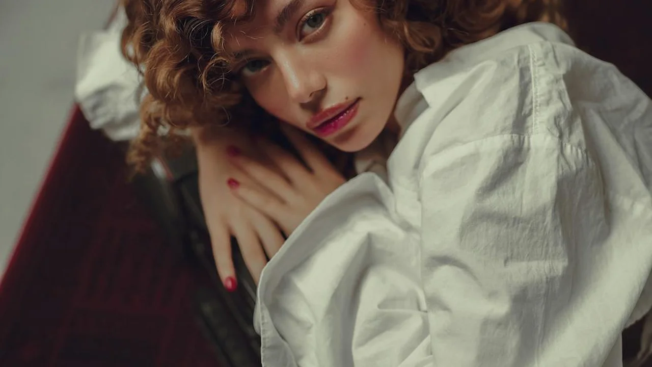 Portrait of a woman with curly hair wearing a white blouse, leaning on a chair.