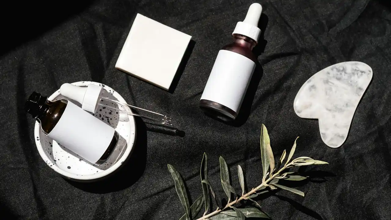 Flat lay of skincare items on a black fabric background, including a dropper bottle, a small white jar with a dropper, a white square sponge, an oval stone, and sprigs of olive leaves.