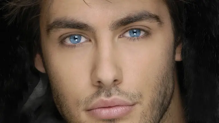 Close-up portrait of a man with blue eyes and light facial stubble wearing a dark hood.