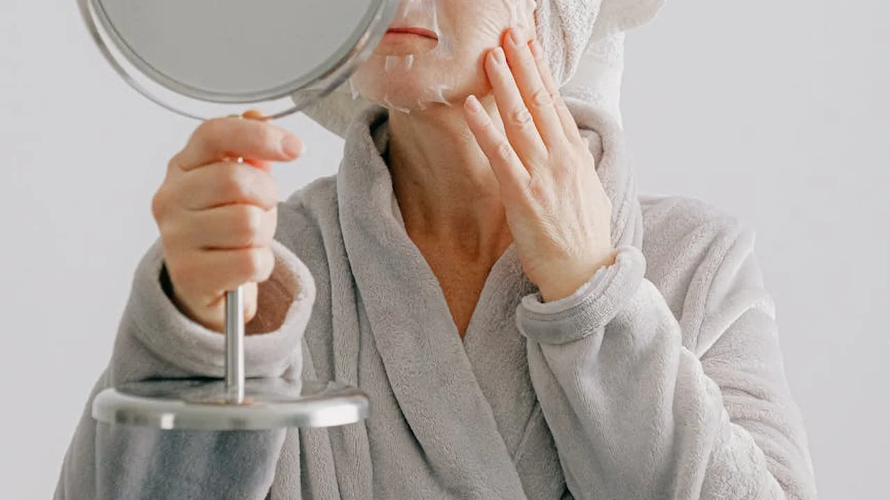 Person in a gray bathrobe applying skincare while looking into a handheld mirror.