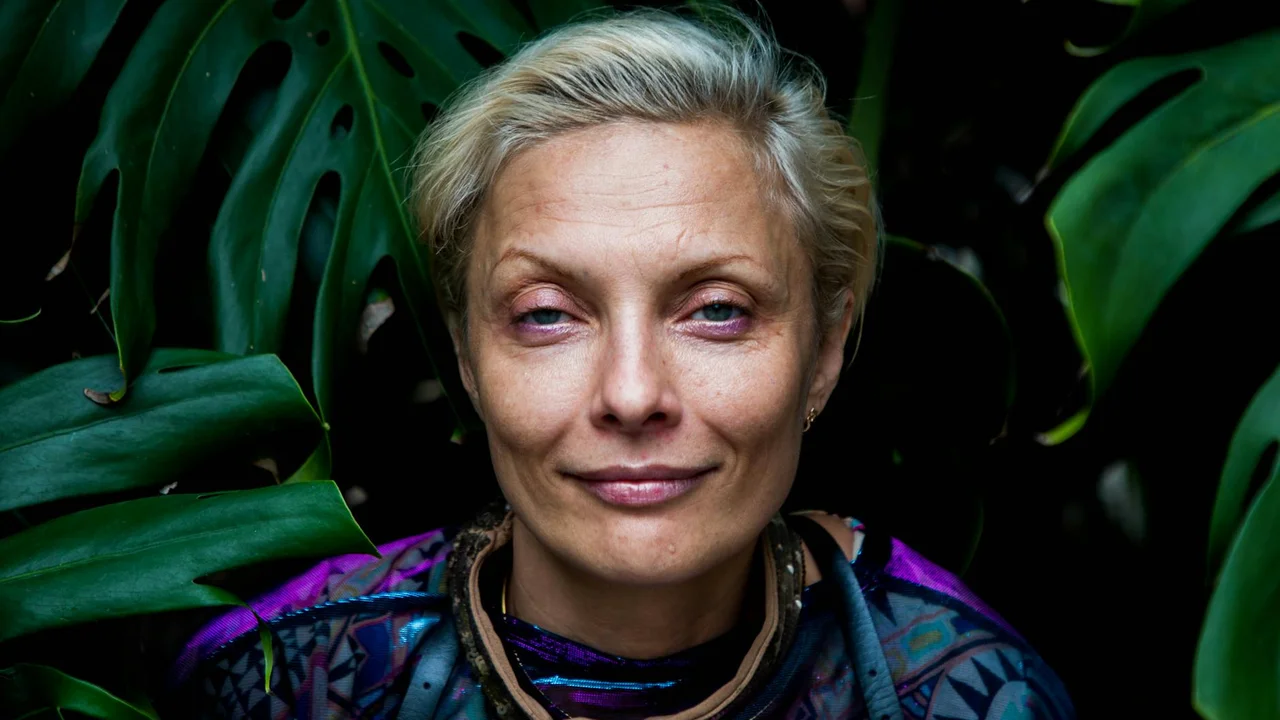 Portrait of a person with short blond hair standing among large tropical green leaves.