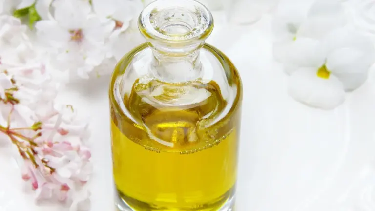 A clear glass bottle of golden facial oil sits on a white surface, surrounded by delicate pink and white flowers.