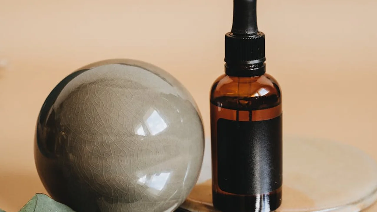 A brown glass dropper bottle sits beside a large glossy spherical oil droplet on a warm beige background, illustrating oil behavior and separation.