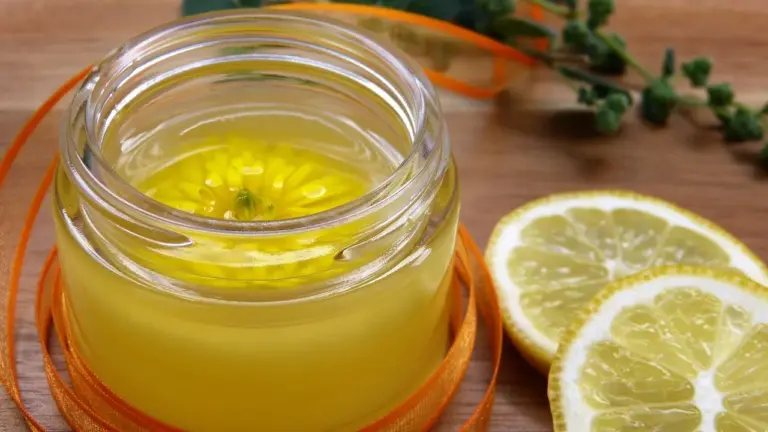 Glass jar of yellow infused oil with lemon slices and fresh herbs on a wooden surface, illustrating at-home botanical oil preparation.