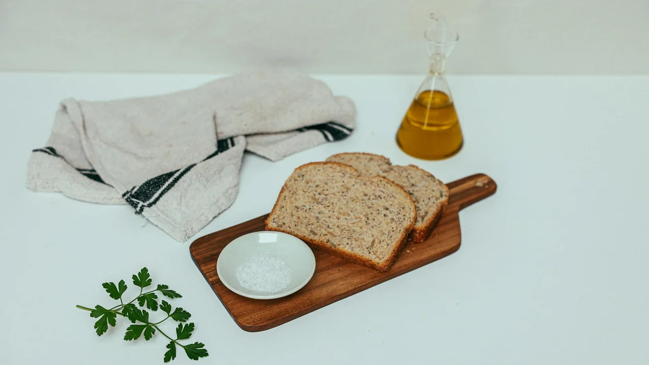Sliced bread on a wooden cutting board with a small white bowl, a bottle of olive oil, a towel, and a sprig of parsley on a white surface.