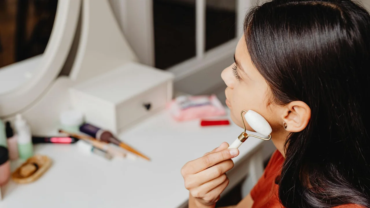 A woman applies skincare using a facial tool at a tidy vanity with skincare products in the background.