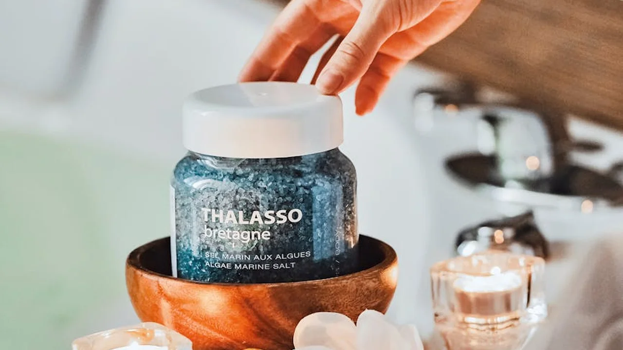 A hand reaching to adjust a jar of sea salt labeled Thalasso Bretagne sitting in a copper bowl, with a lit candle in the background.