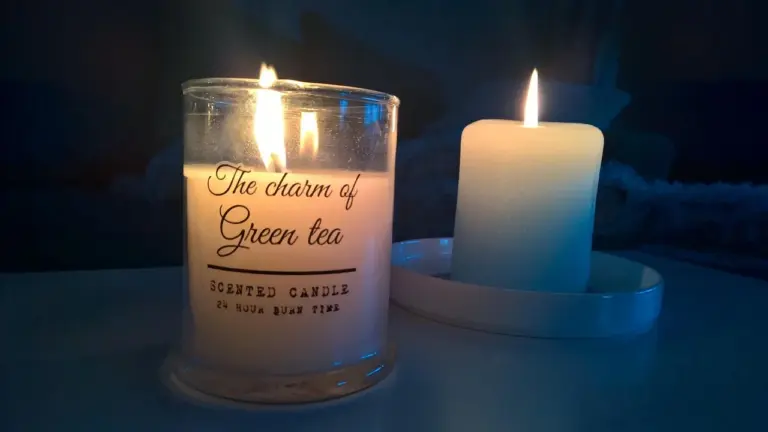 Two lit candles: a glass jar candle labeled 'The charm of Green tea' and a white pillar candle on a blue plate, creating a cozy candle-making mood.