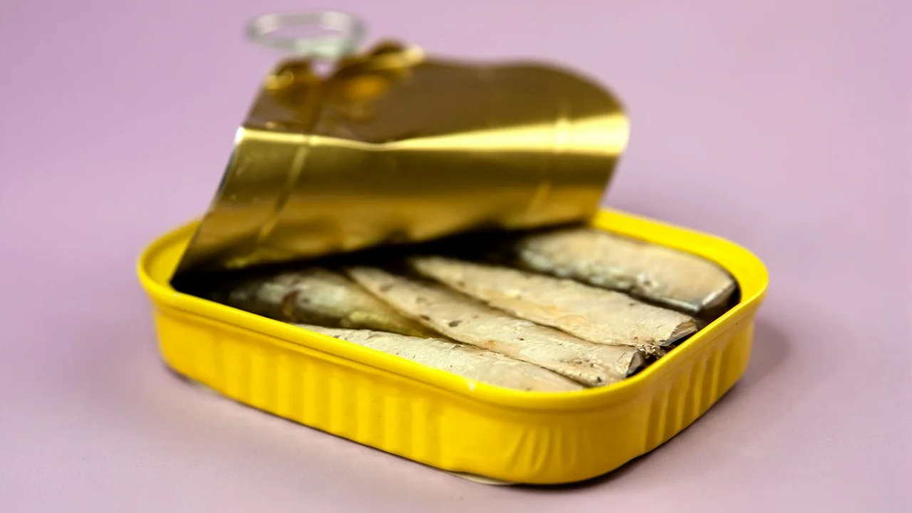 Canned fish in a bright yellow tin with the lid partially opened on a pink background.