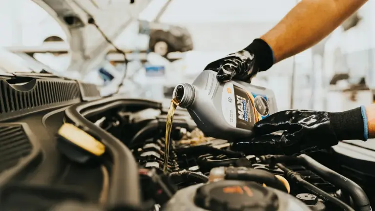 Gloved hands pouring motor oil into a car engine with an oil container visible.