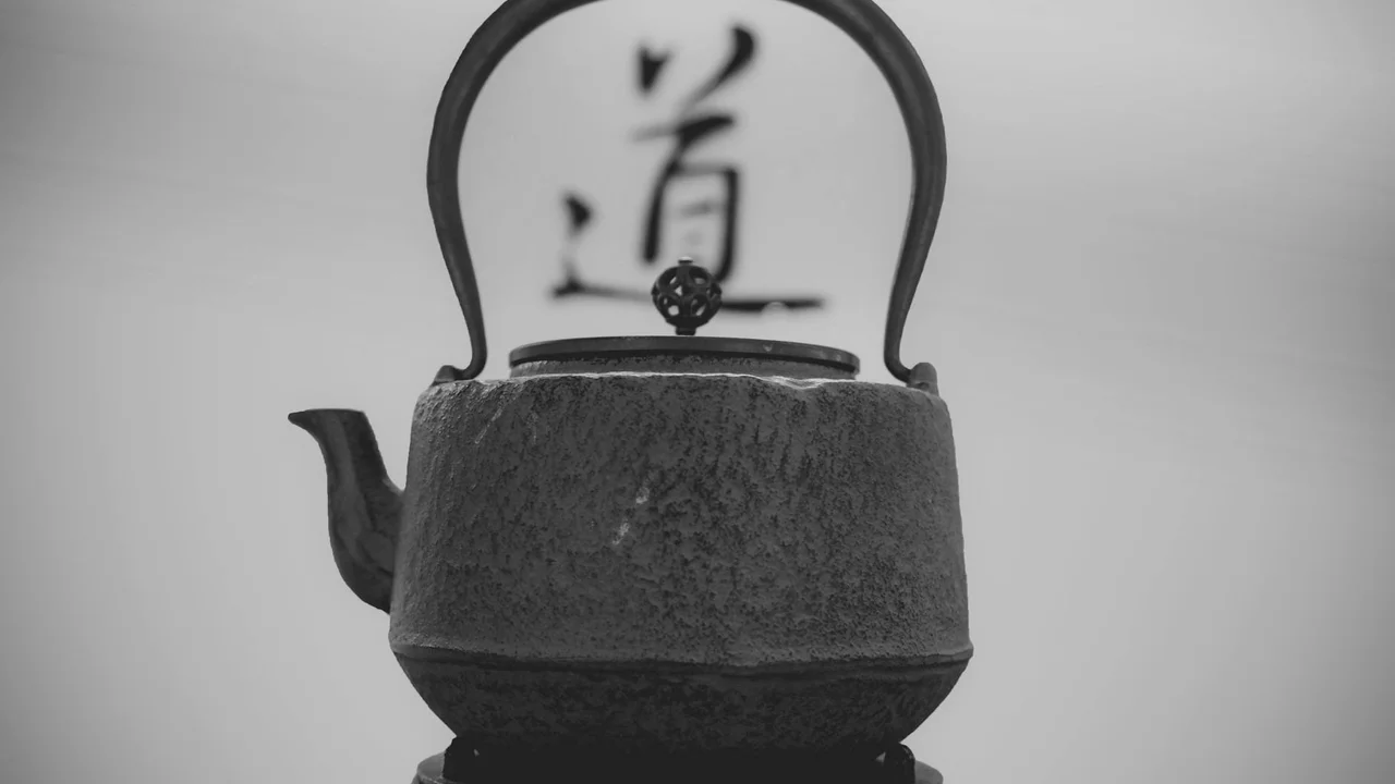 Grayscale image of a black cast iron teapot with a curved handle, set against a soft background featuring a large calligraphic character in the distance