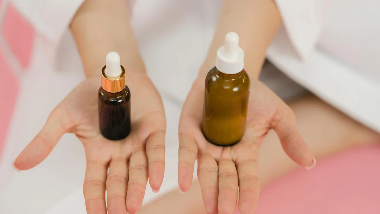Two dropper bottles (one dark brown, one amber) held in open palms against a soft pink background.
