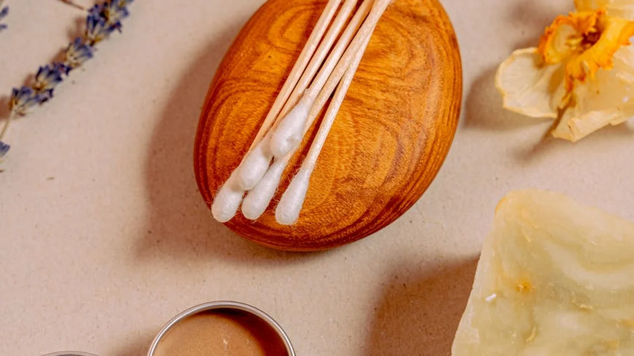 Flat lay of a skincare scene: a wooden bowl holding cotton swabs, with lavender sprigs and a yellow flower on a light beige surface.