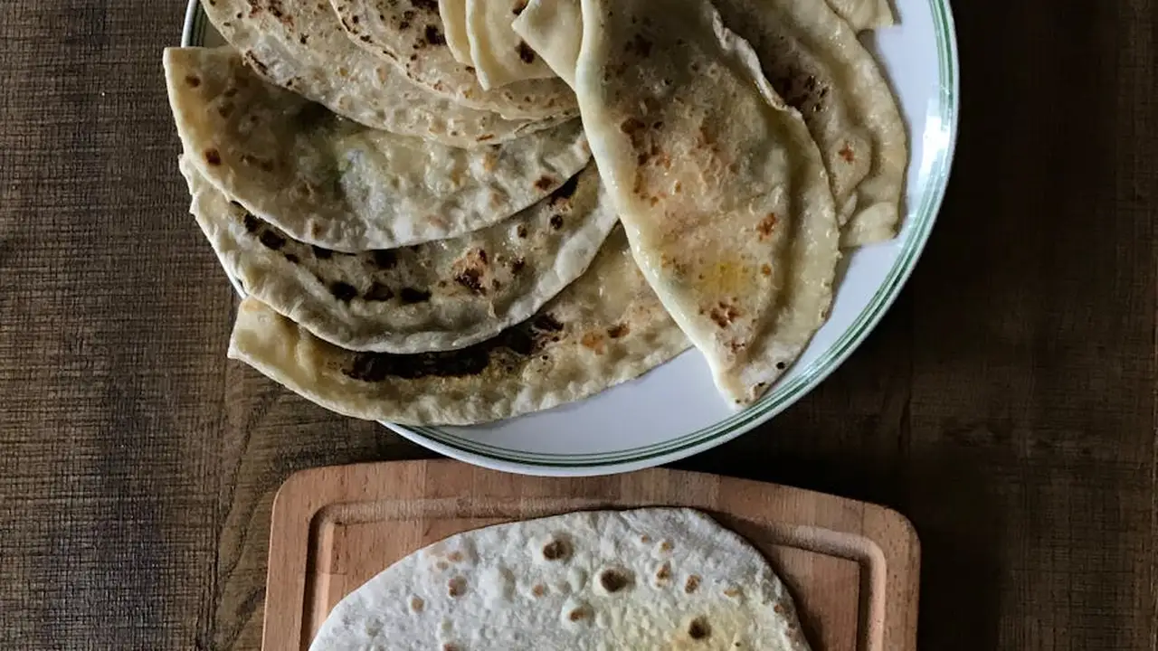 Stack of round, lightly browned chapatis layered on a white plate with a blue rim, set on a dark wooden surface.