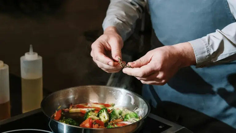 Chef in a blue apron holds a small truffle over a pan of vegetables sizzling on a stove.
