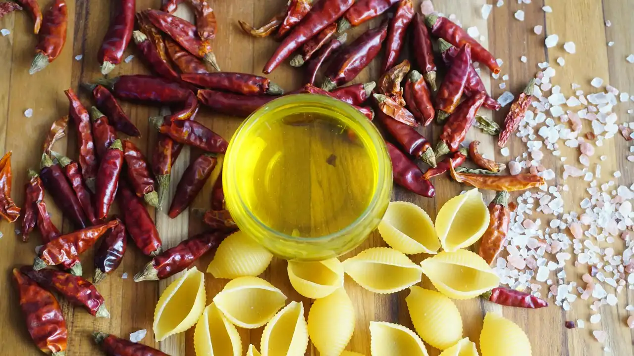 Dried red chili peppers, a small glass bowl of yellow chili oil, and conchiglie pasta topped with sea salt on a wooden surface