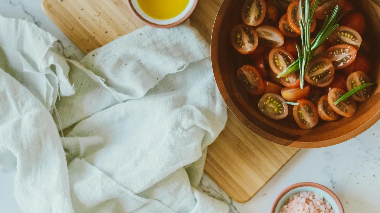 Top-down view of a wooden surface with a bowl of halved cherry tomatoes, a small dish of olive oil, and a white linen cloth, illustrating ingredients for cooking with olive oil.