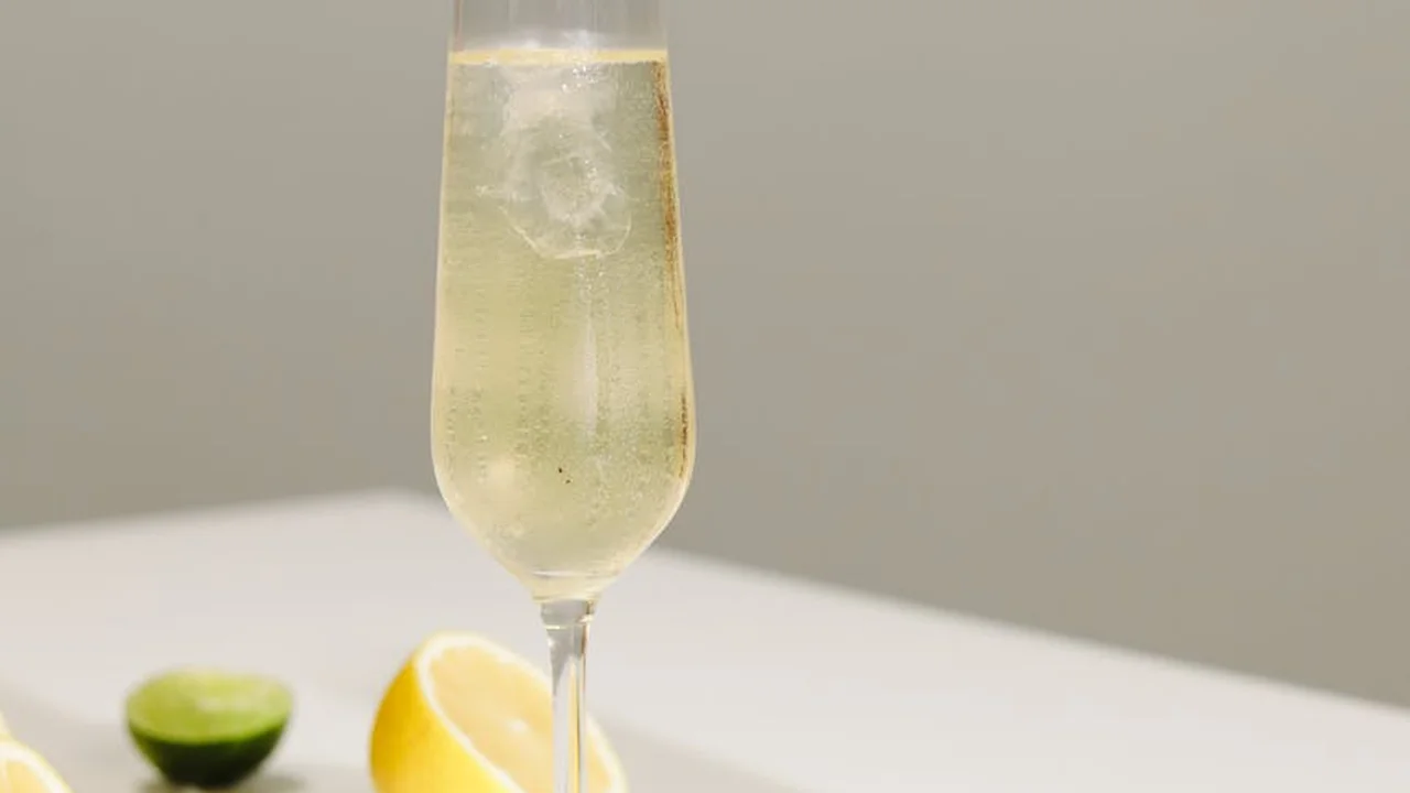 A tall champagne flute with a pale sparkling beverage sits on a light surface, with lime wedges blurred in the background, hinting at fresh citrus aromas.