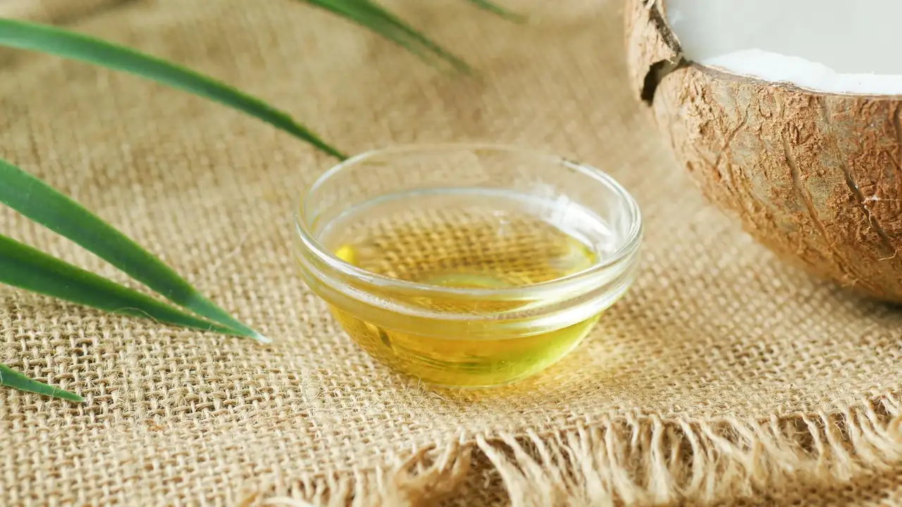 Glass bowl filled with coconut oil on a burlap surface, with a halved coconut and green leaves nearby.