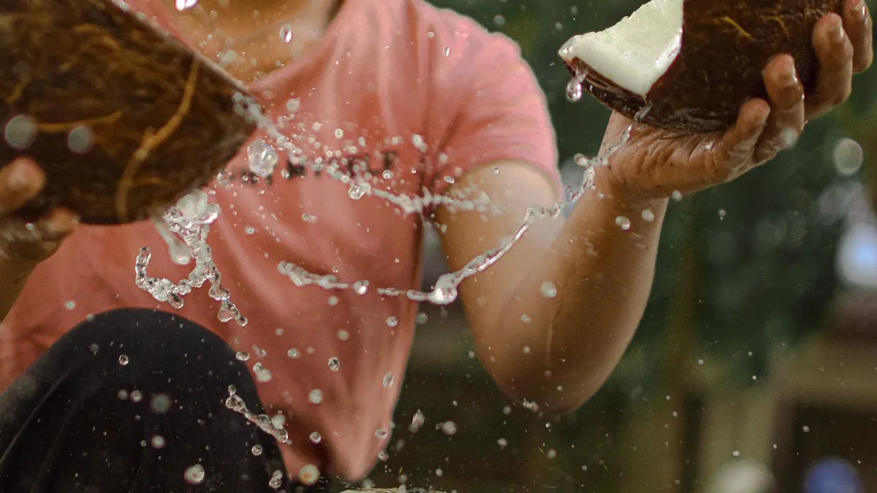 A person in a pink shirt cracks open coconuts with water splashing around their hands, illustrating coconut oil preparation.