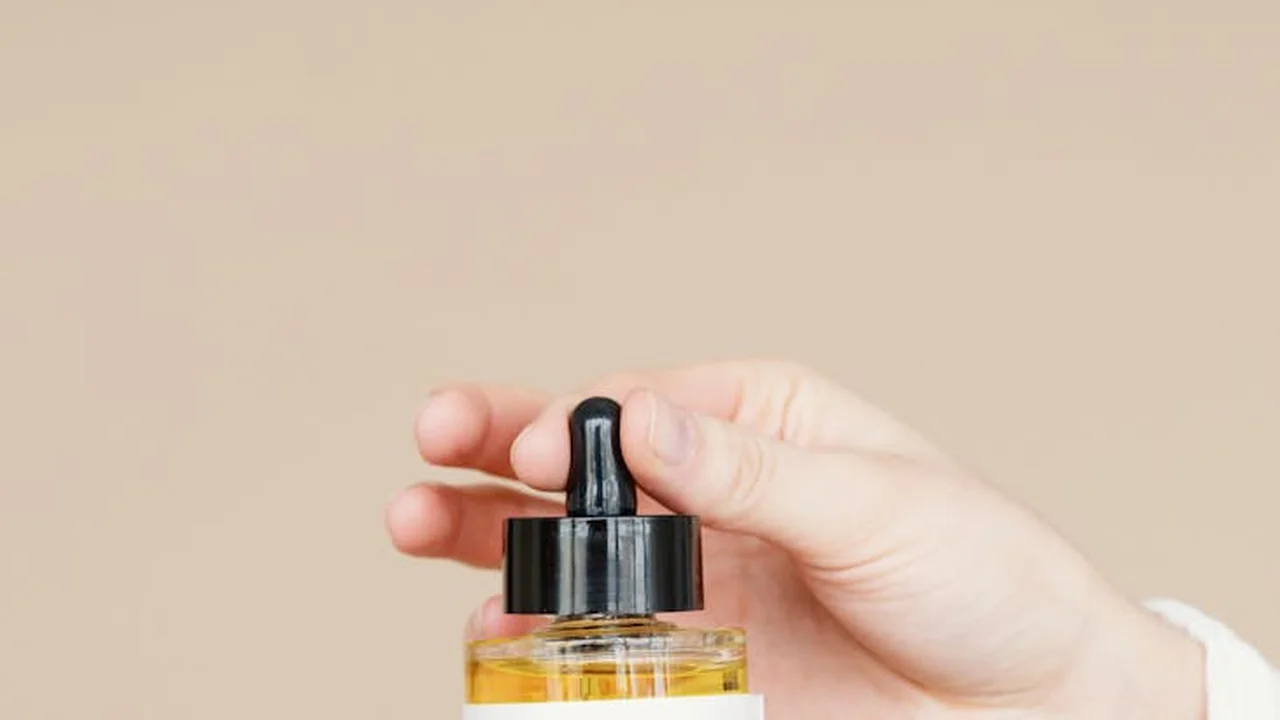 Amber glass dropper bottle filled with golden coconut oil being held by a hand against a neutral beige background.