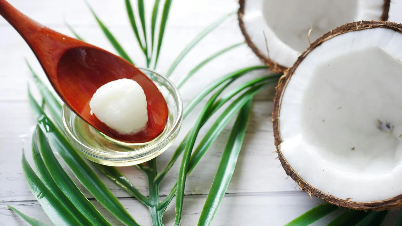 A spoon with coconut oil over green palm leaves and fresh coconut halves on a white wooden surface, illustrating coconut oil as a skin care product for dogs.