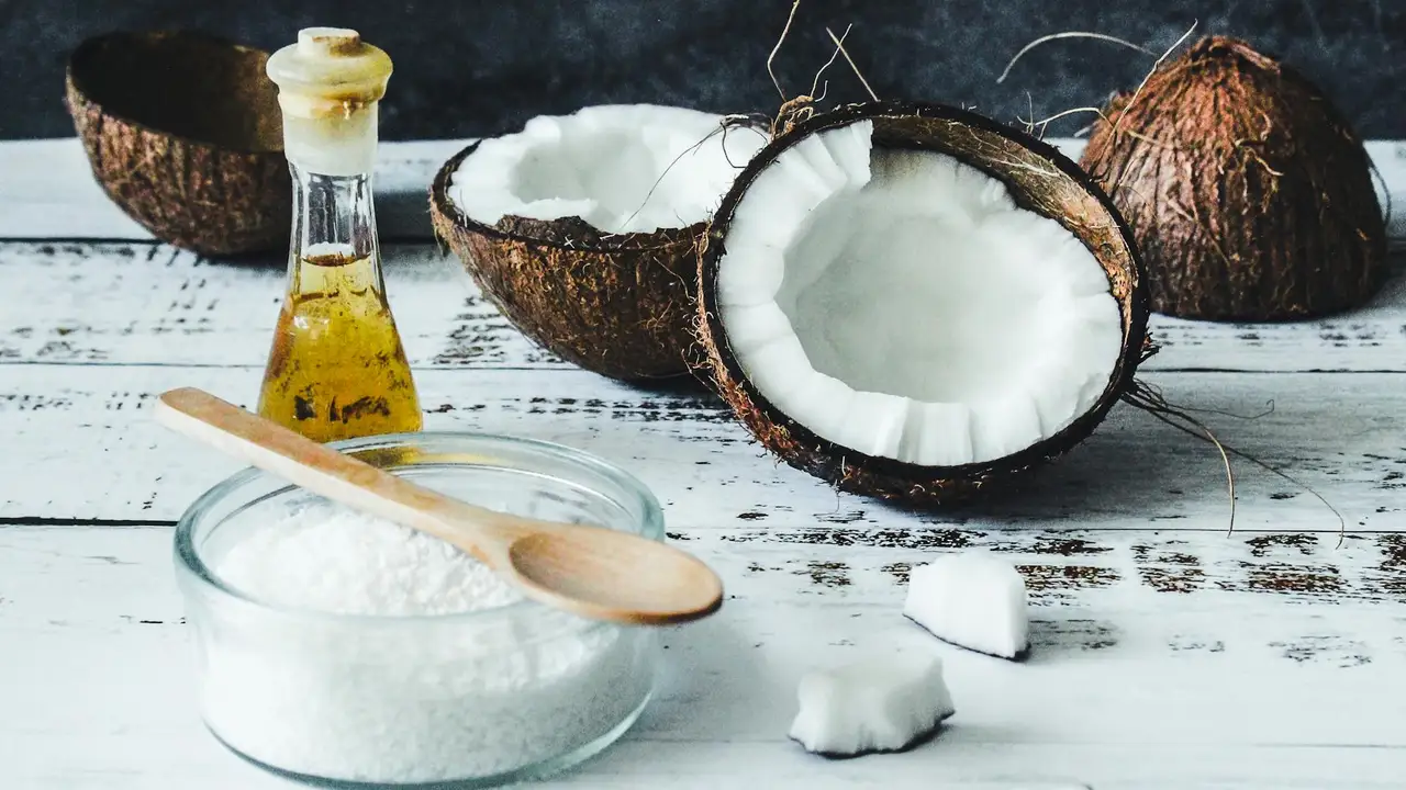 Coconut halves, a bottle of coconut oil, a wooden spoon, and a bowl of white coconut powder on a light wooden surface.