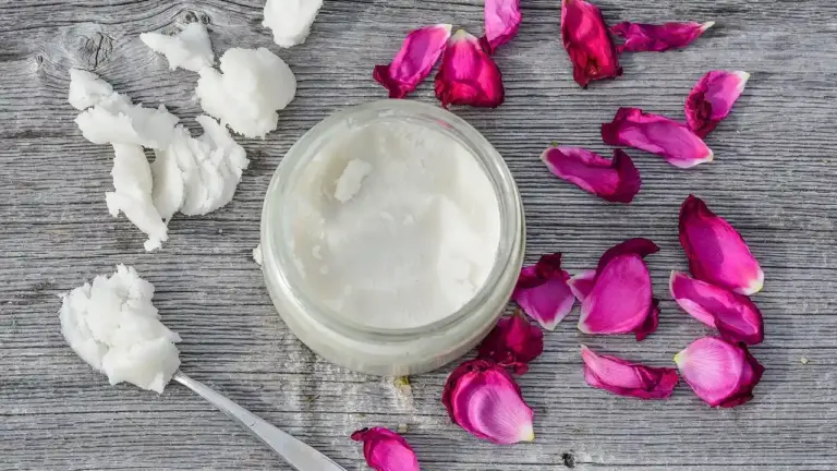 A jar of coconut oil on a wooden surface with scattered coconut pieces and pink rose petals.