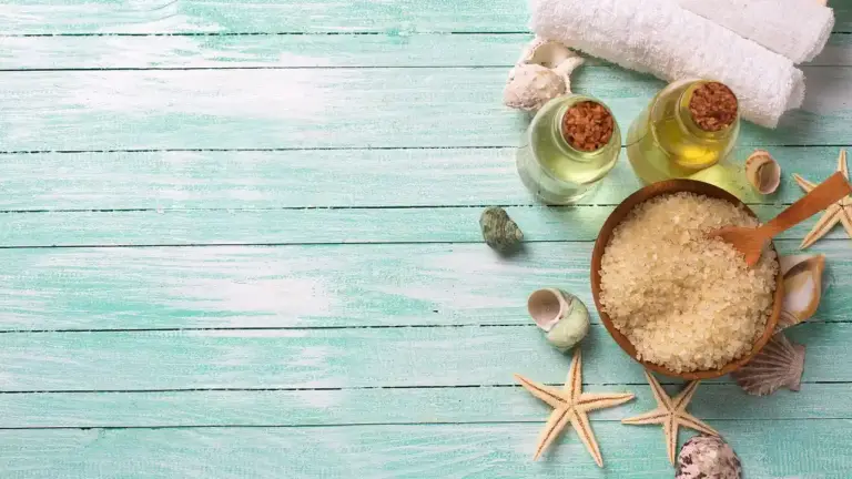 Spa setup with coconut oil bottles, a wooden scoop of sugar, seashells and starfish on a turquoise wooden surface