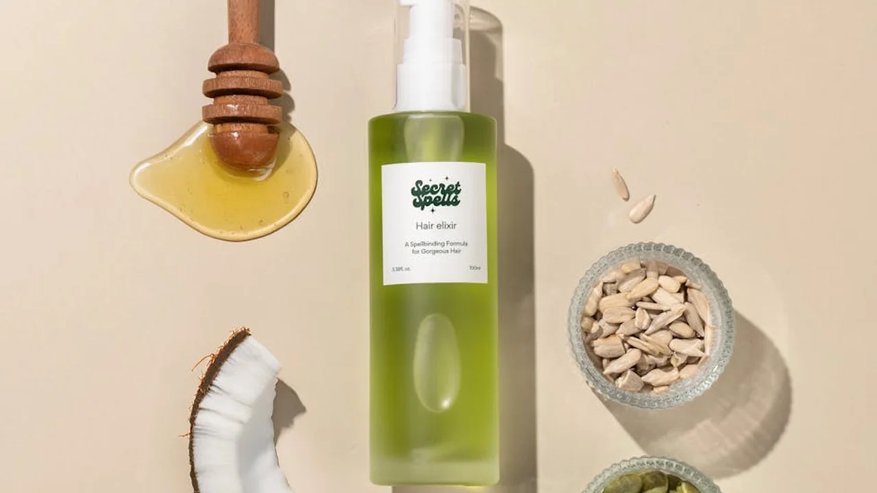 A green bottle of hair oil on a beige background, with a wooden honey dipper and a small bowl of seeds nearby.