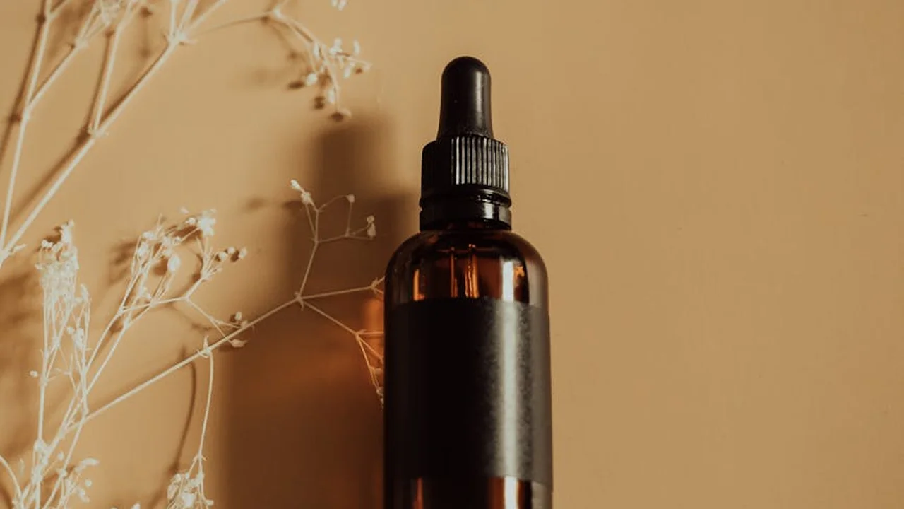 Amber glass dropper bottle on a warm beige background with dried botanical stems nearby, evoking cold-pressed essential oils