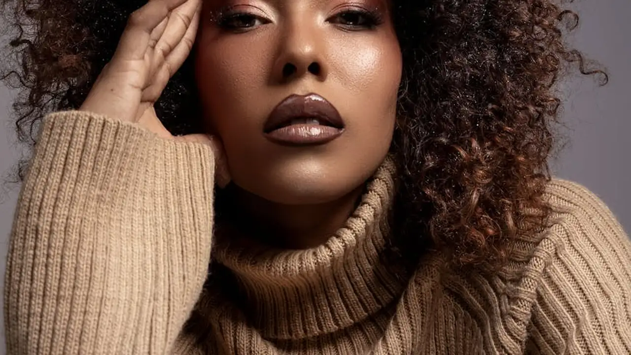 Close-up portrait of a woman with curly hair wearing a beige knit turtleneck, touching her temple.