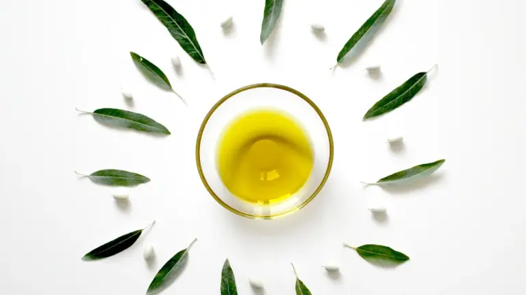 Top-down image of a small glass bowl filled with olive oil, surrounded by green leaves on a white background.