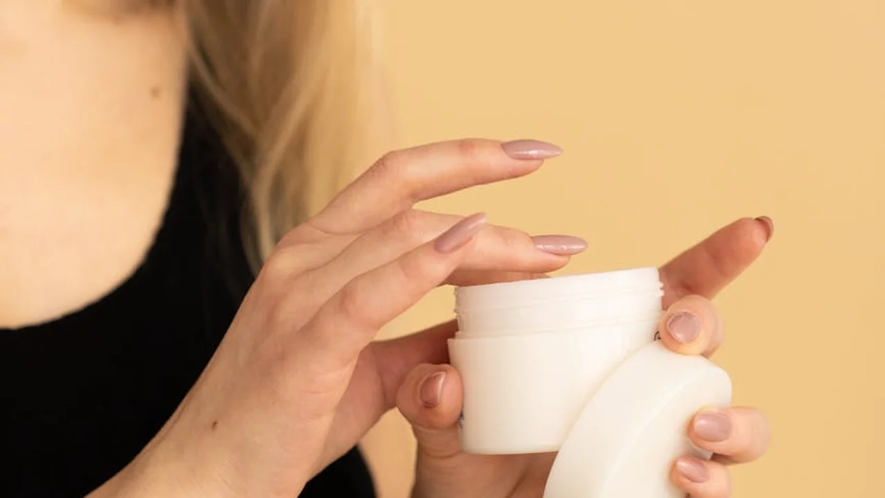 Close-up of hands opening a small white cosmetic jar against a warm beige background, symbolizing checking ingredient labels for non-comedogenic properties.