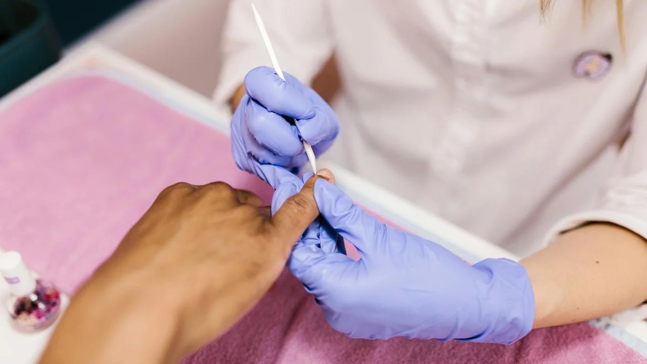 A nail technician wearing purple gloves applies cuticle oil to a client’s cuticles during a manicure.