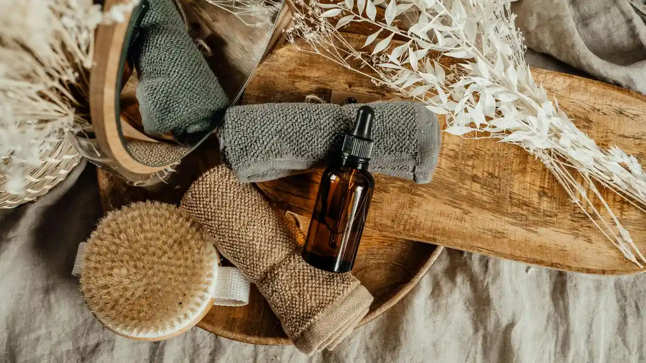 Brown glass essential oil bottle with dropper on a wooden surface, surrounded by rolled towels, a brush, and dried grasses.