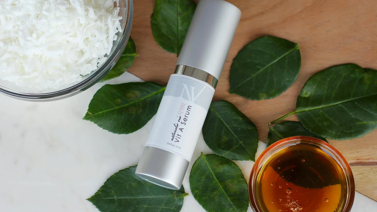 Bottle of coconut oil with shredded coconut and green leaves on a wooden surface, and a small bowl of oil nearby