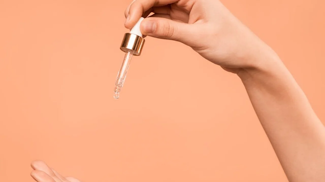 Close-up of a hand holding a dropper releasing liquid against a peach-colored background