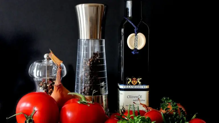 Olive oil bottle, pepper grinder, glass container, and fresh red tomatoes arranged on a dark surface, illustrating ingredients for cooking eggs.