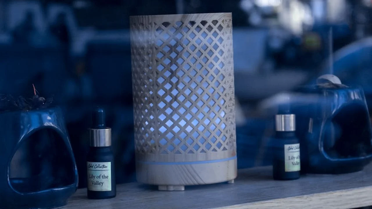 Blue-toned image of a cylindrical essential oil diffuser with a lattice pattern, flanked by small blue oil bottles on a dark wooden surface.