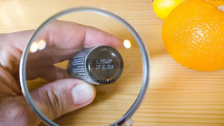 Close-up of a hand holding a small essential oil bottle under a magnifying glass with an orange on a wooden surface.