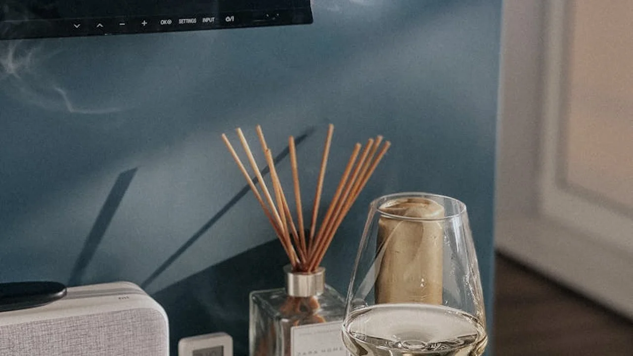 A reed diffuser with wooden sticks in a glass bottle sits on a surface beside a lit candle and a laptop against a blue wall.